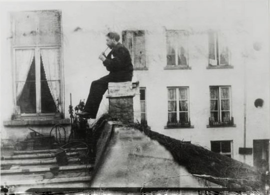 James Ensor jouant de la flûte assis sur une cheminée, mais il tient le flûte entre les lèvres. Photographie anonyme de 1885.